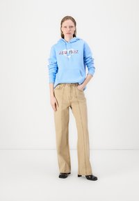 Světle modrá mikina s grafikou textu "NEW YORK", kombinovaná s béžovými kalhotami s širokými nohavicemi. Černé boty dotvářejí outfit. Jednoduché bílé pozadí.