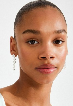 Garas sudraba krāsas auskari ar apaļām un ovālām saitēm, kas atstaro gaismu, valkāti uz personas pret gaišu fonu.