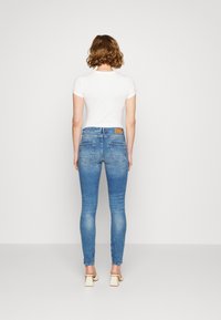 Kvinna med kort lockigt hår iförd en vit kortärmad topp, blå skinny jeans och krämfärgade klacksandaler, stående mot en enkel vägg.
