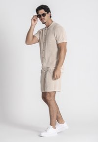 Beige gebreide shirt met korte mouwen en knopen, gecombineerd met bijpassende shorts, en witte sneakers en een zonnebril, tegen een effen achtergrond.