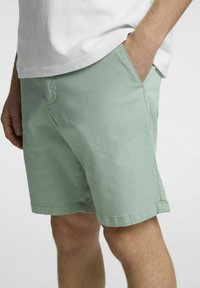 Světlé zelené bavlněné šortky s bočními kapsami, rovný střih a ohrnutý lem. Materiál má hladkou strukturu a minimalistický design.