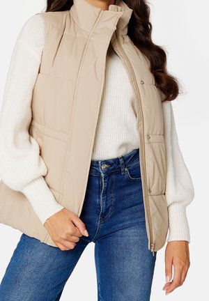 Woman wearing beige padded vest over white knit sweater and blue denim jeans, hand adjusting vest zipper on white background.