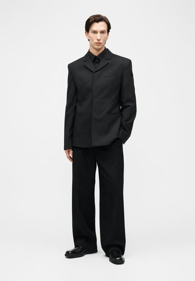 Homme debout portant un blazer noir sur mesure, un pantalon noir à jambes larges, des chaussures habillées noires et une chemise noire à boutons, sur fond blanc uni.