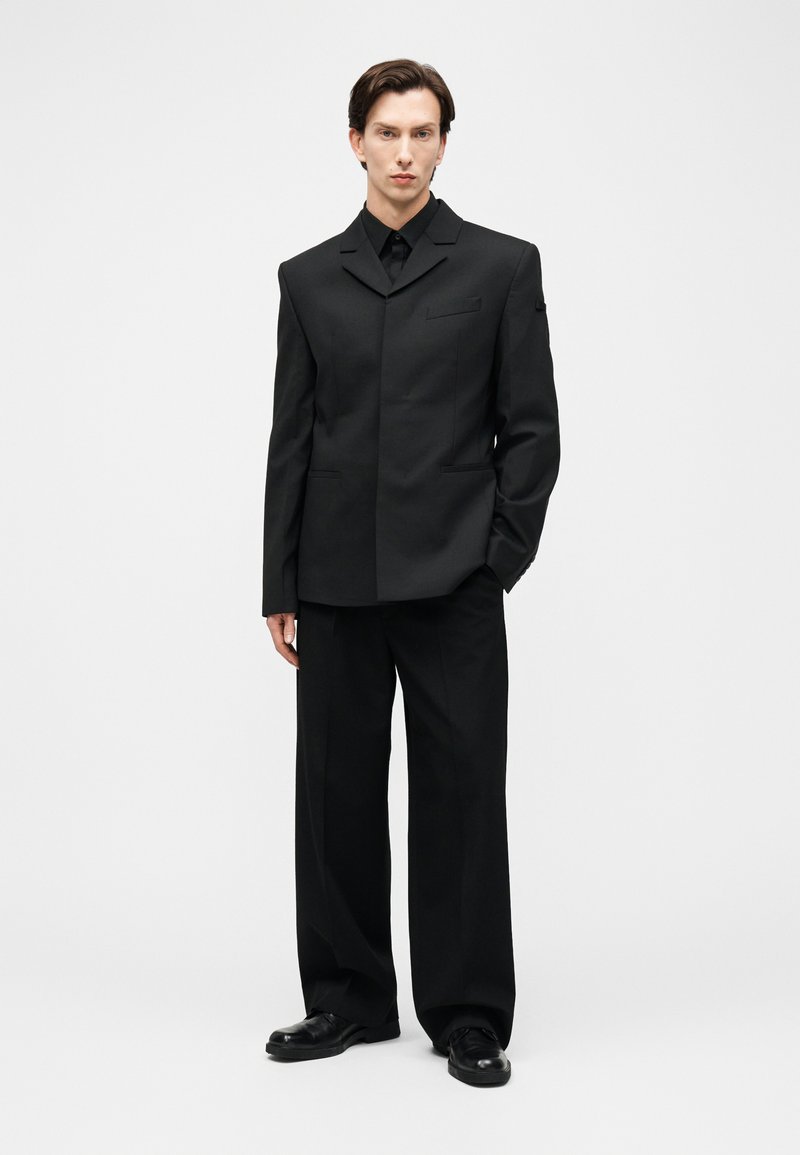 Mand stående iført skræddersyet sort blazer, sorte bukser med vidde, sorte pindestøvler og sort skjorte med knapper mod en ensfarvet hvid baggrund.