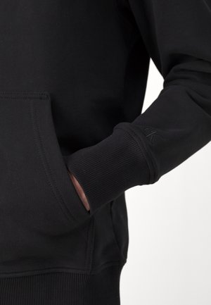 Person trägt einen schwarzen Pullover mit gerippten Bündchen, die Hand in der vorderen Tasche und ein dezentes "CK"-Logo auf dem Ärmelbündchen gestickt.