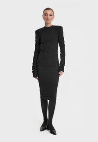 Czarna sukienka z długim rękawem o dopasowanym kroju, z marszczonymi rękawami i wysokim dekoltem. Stylizowana z czarnymi rajstopami i czółenkami z ostrym noskiem.