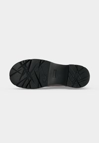 Semelle de chaussure noire avec des motifs de crampons profonds et inclinés et un talon légèrement surélevé, vue du dessous sur un fond clair uni.