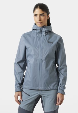 Mujer con chaqueta impermeable azul con capucha y pantalones a juego, de pie con las manos relajadas, mirando ligeramente hacia un lado.
