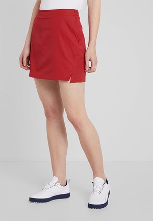 Femme portant une jupe rouge avec une fente latérale, des baskets blanches et un haut blanc, debout devant un fond gris clair uni.