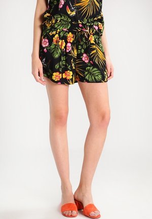 Kvinde iført sorte shorts med tropisk blomsterprint og orange åbne, lave hælede slippers, stående mod en ensfarvet hvid baggrund.