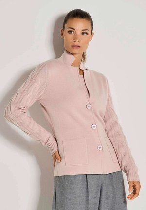 Vrouw in een lichtroze knoopcardigan met gestructureerde kabelgebreide mouwen en grijze broek, staand tegen een lichte muur.