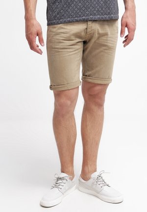 Khaaki spodenki z podwiniętymi nogawkami, wykonane z strukturalnej tkaniny. Noszone z białymi sneakersami i wzorzystą ciemnoszarym koszulą.