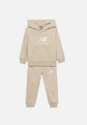 Μπεζ παιδικό φούτερ New Balance με μπροστινή τσέπη και αντίστοιχο παντελόνι φόρμας, και τα δύο με λευκά λογότυπα New Balance.