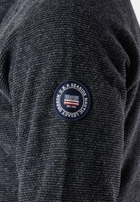 Langarmshirt mit navy- und grauen Streifen aus strukturiertem Stoff, mit einem runden marineblauen Patch am Ärmel, auf dem "Seaside Nautical Legacy" steht.