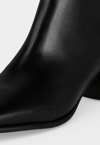 Gros plan d'une bottine en cuir noir à bout pointu et talon bloc bas sur fond blanc.