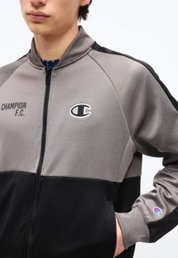 Νέος άνθρωπος που φοράει ζακέτα Champion F.C. με φερμουάρ σε γκρι και μαύρο χρώμα, με λογότυπα στο στήθος και στο μανίκι, το χέρι στην τσέπη.