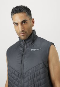 Sleeveless, dark gray vest made of padded material with a smooth texture. Features a front zipper and logos "BARÇA" and "Nike" in white.