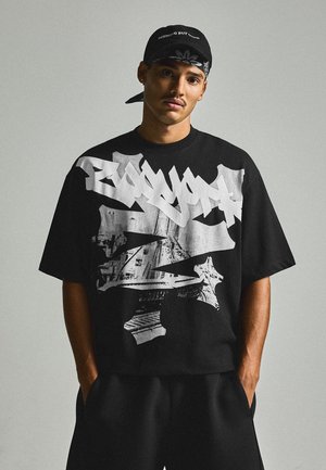 Jeune homme portant un t-shirt noir à motif graphique, une casquette noire avec un texte et un short noir, debout les mains dans les poches devant un fond uni.