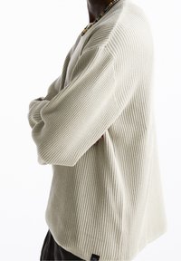 Beiger, gerippter Strickpullover mit langen Ärmeln und lockerer Passform. Verfügt über ein kleines Label am Saum. Texturiertes, weiches Material.