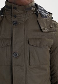 Veste vert olive avec un col montant, plusieurs poches à l'avant, fermetures par boutons, et accents noirs sur les épaules et la capuche. Matériau texturé.