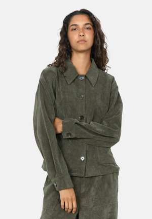 Jeune femme aux longs cheveux bouclés porte une veste boutonnée en velours côtelé vert olive et un pantalon assorti, debout avec un bras croisé.