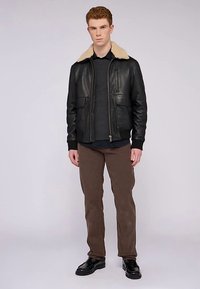 Černá kožená bomber bunda s béžovým kožíškovým límcem, zapínáním na zip a dvěma předními kapsami, ve spojení s tmavým svetrem a hnědými kalhotami.