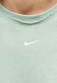 Κοντινό πλάνο ανοιχτοπράσινου υφάσματος με λευκό λογότυπο Nike swoosh κάτω από ένα φθαρμένο λαιμόκοψη σε ένδυμα.