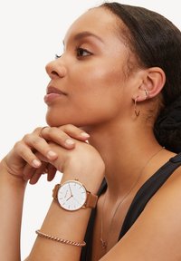 Orologio in pelle marrone con quadrante bianco e dettagli in oro rosa, indossato a un polso. Include un bracciale in catena argento e orecchini a cerchio.