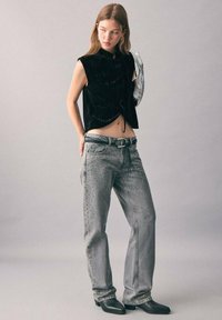 Čierny sametový cropped top s vysokým golierom, v kombinácii s šedými denimovými nohavicami s cvočkovými detailmi a dekoratívnym pásom. Čierne členkové topánky.