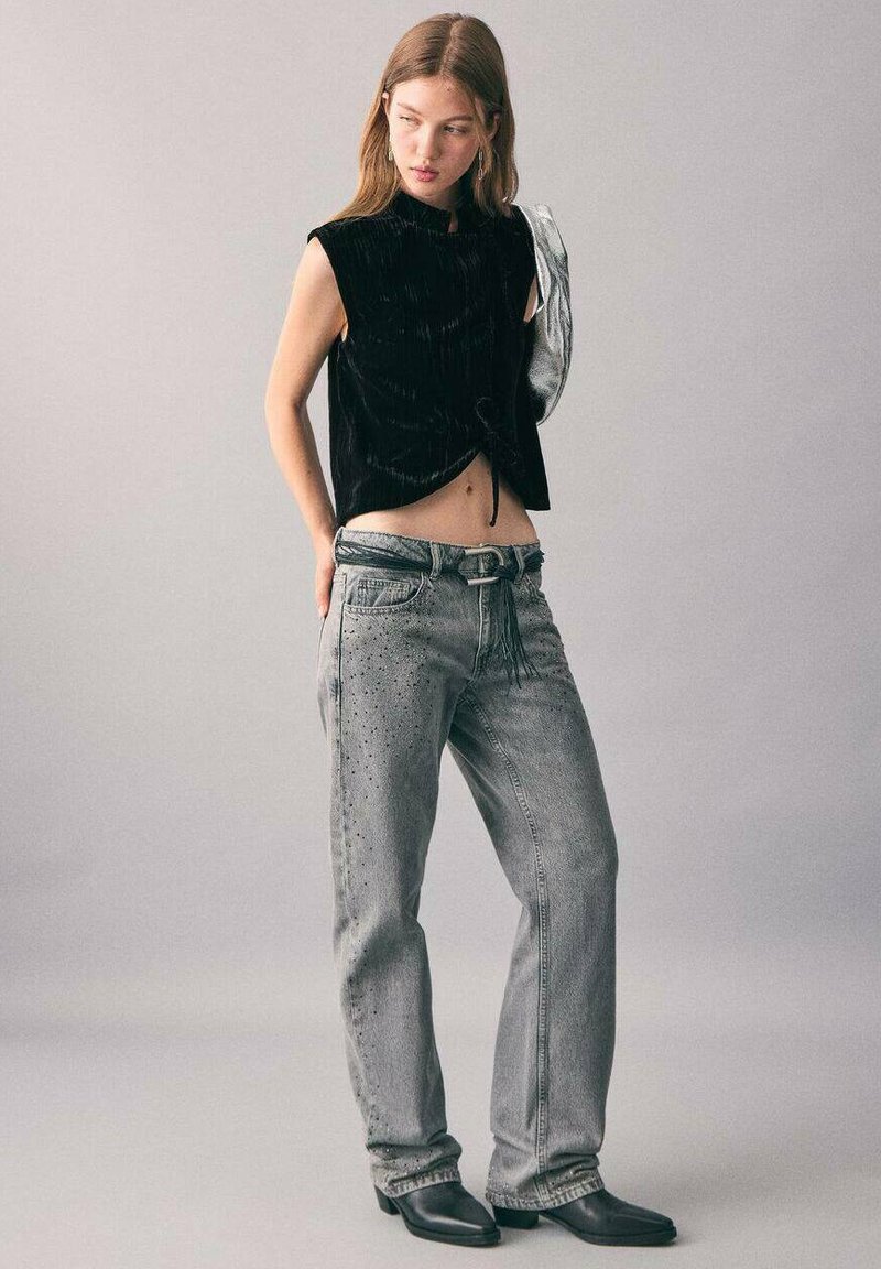 Čierny sametový cropped top s vysokým golierom, v kombinácii s šedými denimovými nohavicami s cvočkovými detailmi a dekoratívnym pásom. Čierne členkové topánky.