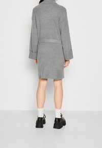 Manteau en mélange de laine gris avec un col montant, des manches longues et un détail noué à la taille. L'ourlet présente une texture subtilement effilochée.