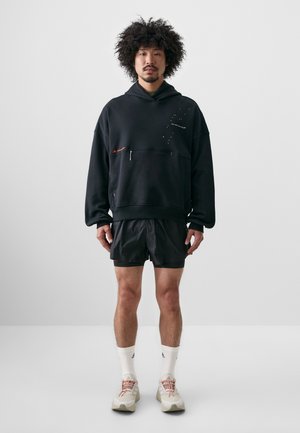 Svart oversized hoodie med en framfickor med dragkedja, med vita och röda accenter. Matcher med svarta shorts, vita strumpor och krämfärgade sneakers.