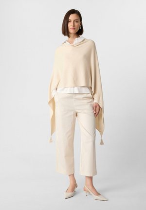 Beige poncho with a soft texture, hooded design, and tassel accents. Worn over a white blouse and paired with cream trousers and light heels.