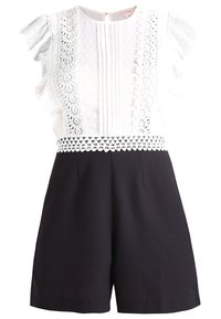 Blouse blanche avec accents en dentelle et plis, associée à un short noir. Manches volantées et détail décoratif à la taille.