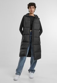 Langer schwarzer Puffer-Westen mit Kapuze, mit einem gesteppten Design, Frontreißverschluss und Seitentaschen, getragen über einem schwarzen Oberteil und blauen Jeans.