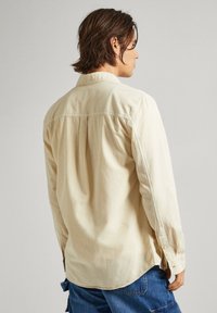 Uomo con capelli castani di lunghezza media che indossa una maglietta a maniche lunghe color avorio e jeans blu, di spalle su uno sfondo semplice.