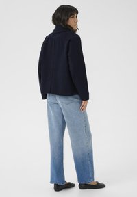 Jūrinės vilnos mišinio striukė su pjautuviniu apykakle, dėžutės formos kirpimu ir užpakaline siūle. Kombinuota su šviesiai mėlynomis aukšto liemens džinsais ir juodais butais.