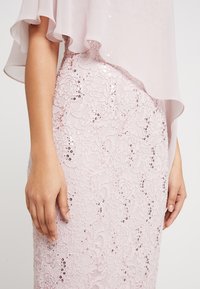 Robe rose clair présentant une texture en dentelle avec des sequins, des manches en chiffon superposées et une silhouette ajustée. Gros plan sur une main sur le tissu.