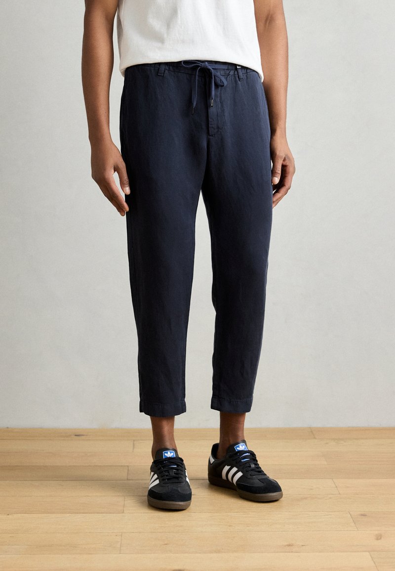 Marineblauwe linnenmixbroek met een elastische tailleband en trekkoord, cropped lengte, gecombineerd met zwarte sneakers met witte strepen.