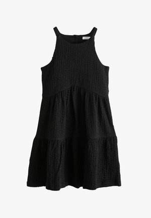 Djevojka s tamnom kovrčavom kosom vezanom u rep nosi crnu haljinu bez rukava od teksturiranog materijala s otvorenim leđima pričvršćena na dva gumba.