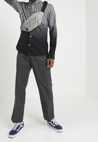 Person som bär en svart-grå gradient rutad skjorta, grå byxor, en grå crossbody-väska och lila sneakers med vita snörningar och sulor.