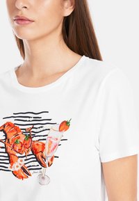 Weißes Baumwoll-T-Shirt mit einem Hummer- und Cocktailsujet in Rot und Pink, akzentuiert mit grünen Perlen, vor schwarzem Streifenmuster.