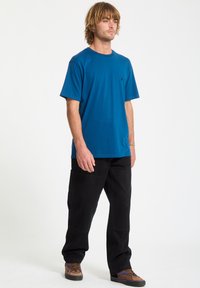 Türkisfarbenes Baumwoll-T-Shirt mit Rundhalsausschnitt, kurzen Ärmeln und einem kleinen Logodetail. Kombiniert mit schwarzen Cargo-Hosen und braunen Sneakers.