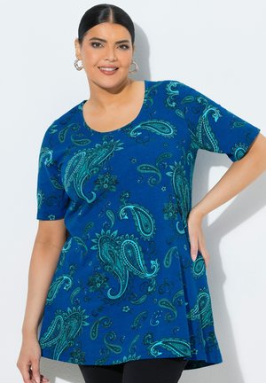 Lächelnde Frau mit dunklen Haaren trägt Creolen und eine blau gemusterte Kurzarm-Tunika mit Paisley-Motiv, posiert mit einer Hand in der Hüfte.