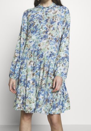 Femme portant une robe à manches longues, longueur genou, avec un motif floral abstrait bleu, vert et marron et une jupe à volants froncée.