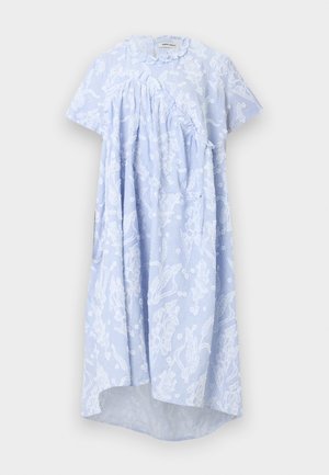 Woman wearing a loose, light blue dress with white floral patterns and ruffles, paired with dark red lace-up flat shoes, standing against a plain background.