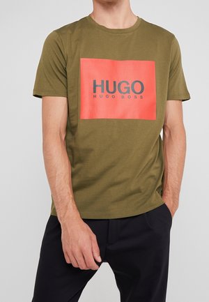 Oliwkowa zielona bawełniana koszulka z prostokątnym czerwonym nadrukiem z napisem „HUGO” w pogrubionym czarnym tekście. Krótkie rękawy i okrągły dekolt.