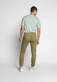 Lysegrønn t-skjorte med korte ermer, kombinert med olivengrønne, smale bukser og hvite joggesko med gule detaljer, sett fra bak.
