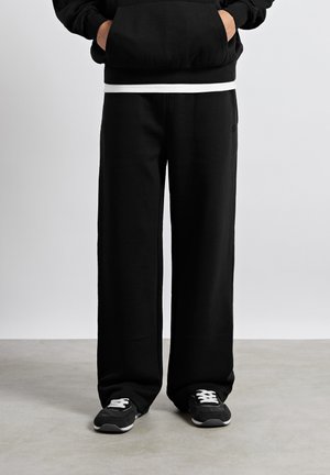 Person iført løse sorte joggingbukser, sorte sneakers med hvide snørebånd og en sort hættetrøje med hænderne i lommerne.