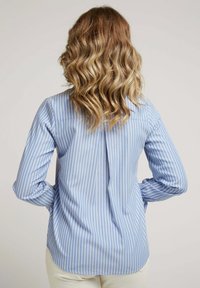 Chemise rayée bleue et blanche à manches longues, avec un pli dans le dos pour plus de facilité et un ourlet courbé. Le tissu semble lisse avec une légère texture.
